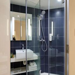 Shower stall with glass doors. Mirror, sink and neatly rolled towels in the background.