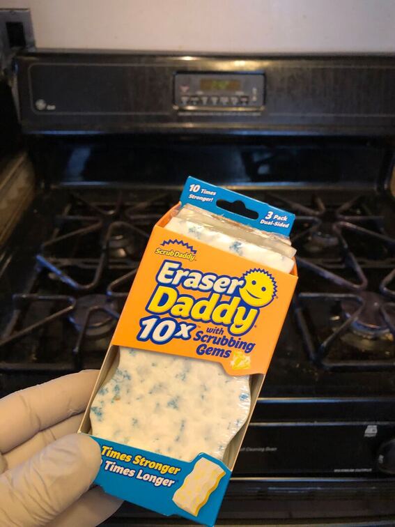 A man's hand hold an unopened three pack of Scrub Daddy brand Eraser Daddy with Scrubbing Gems over a dirty stove.