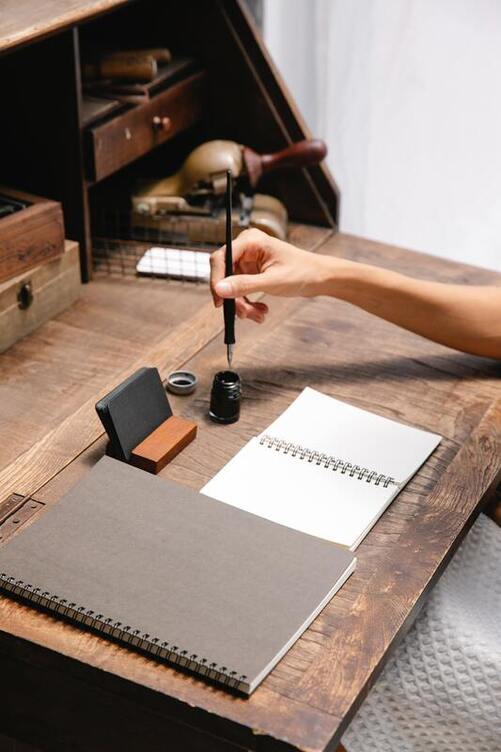 White womon's hand holding a dip pen just above an inkwell. The pen sits on secretary desk with a large notebook, which is closed. A small notebook is opened to a blank page with now lines. 